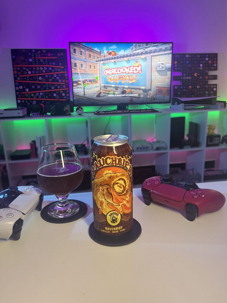 Cerveja escura estilo Rauchbier ao lado de gameplay cooperativo de Overcooked no PS5 com dois controles na mesa.