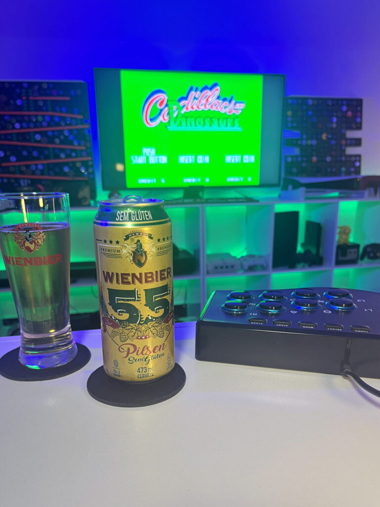 Cerveja Pilsen sem glúten amarelo claro ao lado de Cadillacs and Dinosaurs no Arcade em clima retrô.