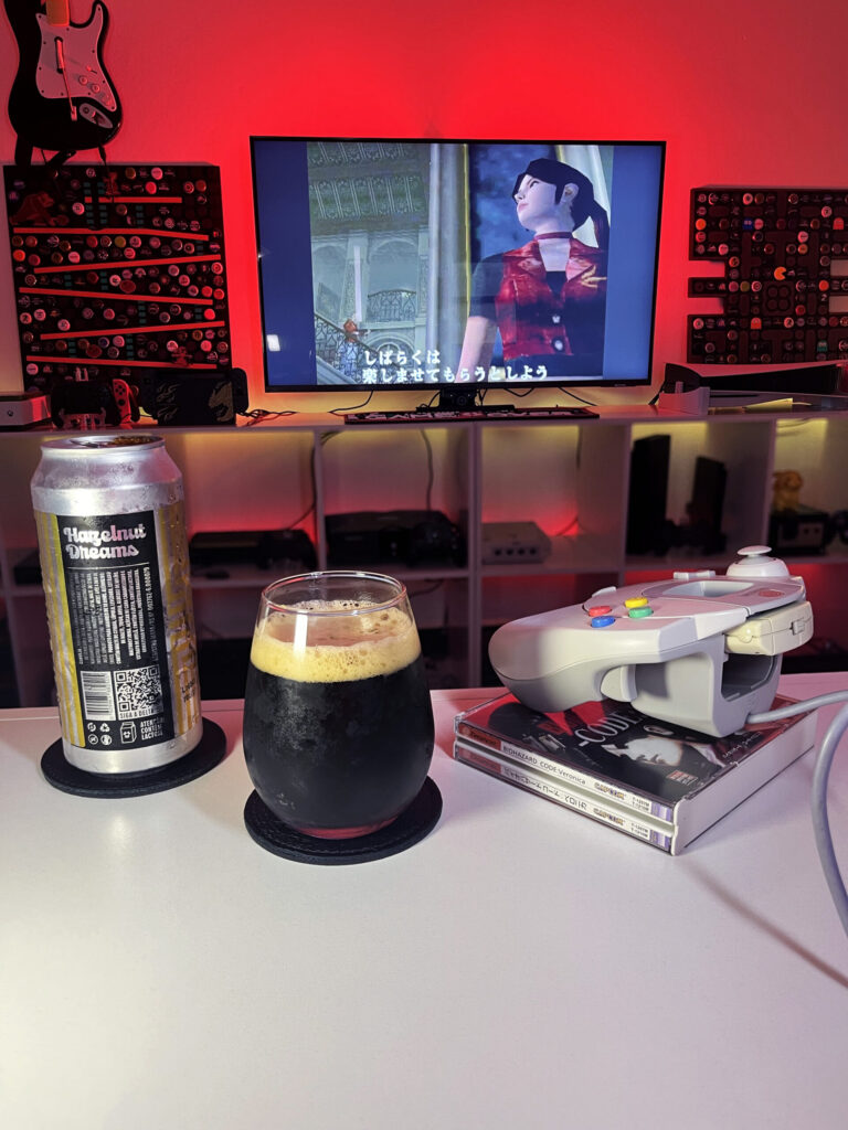Cerveja Hazelnut Dreams negra com espuma marrom clara, ao lado de Biohazard Code: Veronica no Dreamcast.