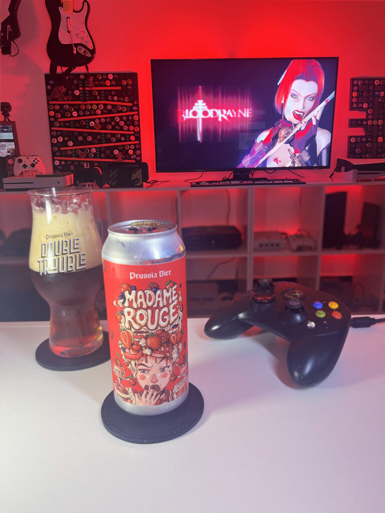 Cerveja Madame Rouge vermelho escuro com espuma marrom clara, ao lado de BloodRayne no Xbox clássico.