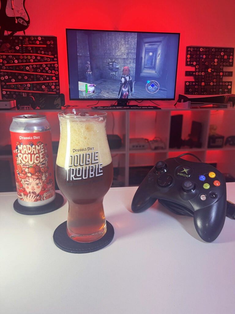 Cerveja Madame Rouge vermelho escuro com espuma marrom clara, ao lado de BloodRayne no Xbox clássico.