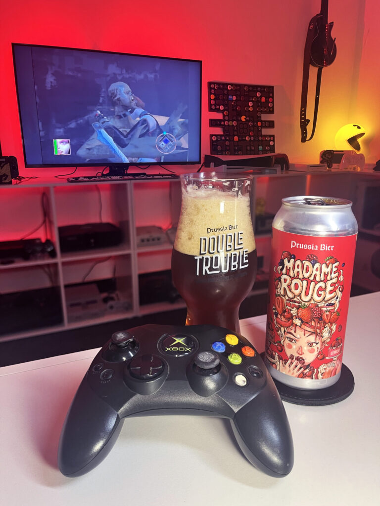 Cerveja Madame Rouge vermelho escuro com espuma marrom clara, ao lado de BloodRayne no Xbox clássico.