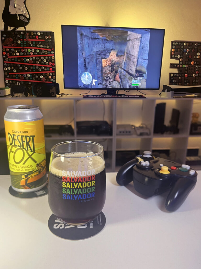 Cerveja Desert Fox cor de carvalho com nuances rubis, ao lado de Call of Duty 2: Big Red One no GameCube.